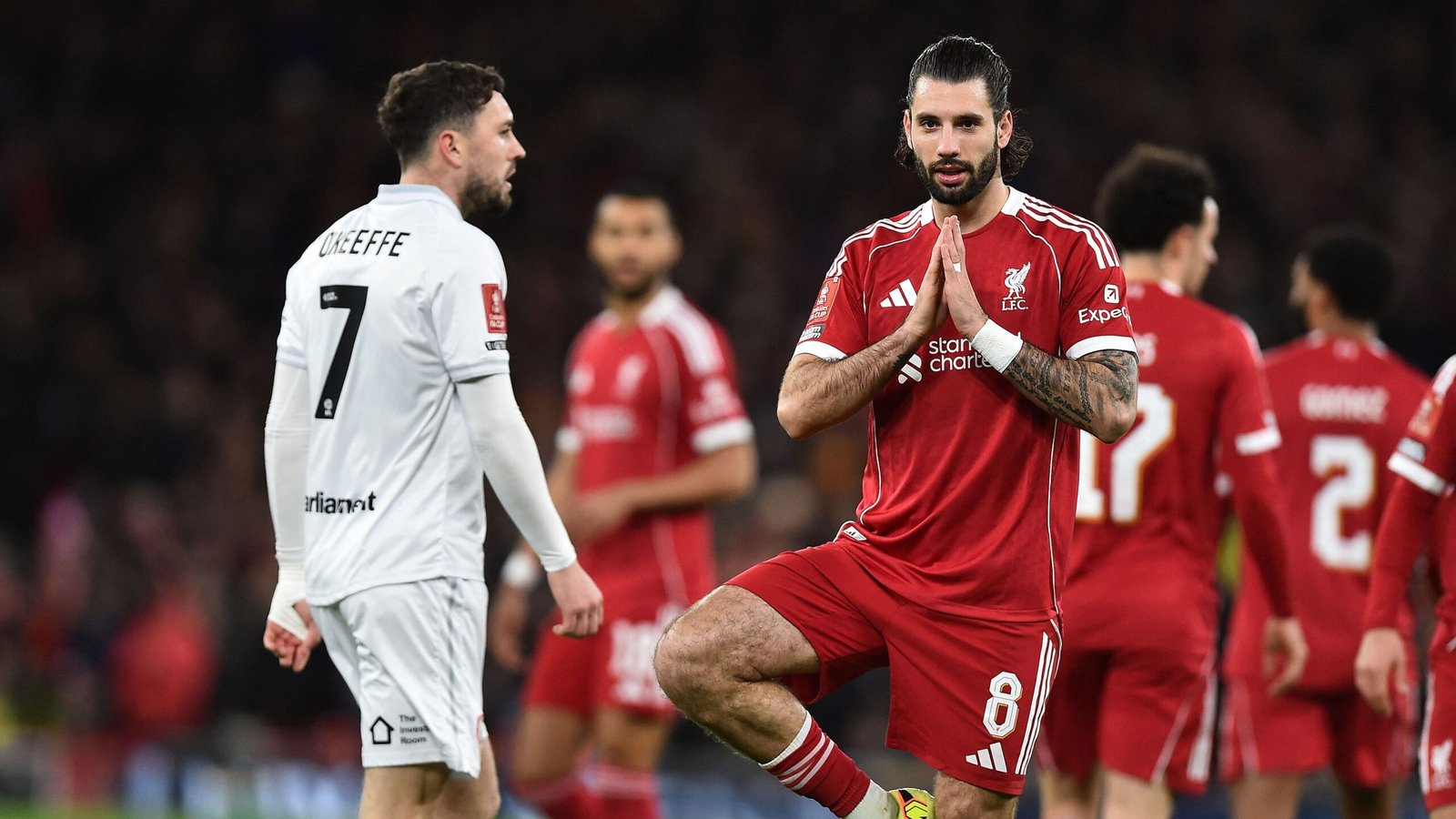 Liverpool's nummer 8 speler knielt in gebed, terwijl nummer 7 van het tegenpartij, O'Keeffe, voorbijloopt. Teamleden zijn op de achtergrond.