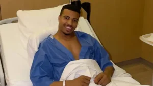 Moroccan footballer in a hospital bed, wearing a blue gown and smiling, showing resilience after a recent medical procedure.