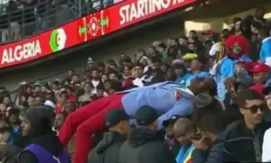Des supporters de football marocains dans un stade, dont un fan allongé de manière ludique sur le côté, au milieu d'une foule animée et de bannières de l'équipe.