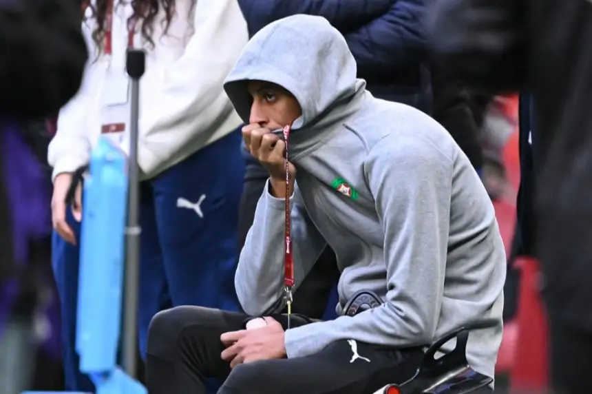 Le joueur marocain est assis pensivement dans un sweat à capuche gris, regardant le match avec attention pendant que ses coéquipiers s'échauffent en arrière-plan.