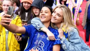 Deux joueuses de football, l'une vêtue du maillot bleu de Chelsea avec une médaille de vainqueur, prenant joyeusement un selfie au milieu de supporters en liesse.