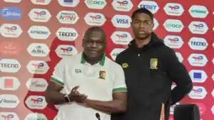 L'entraîneur et le joueur camerounais posent devant un fond de sponsor lors d'une conférence de presse, affichant la fierté de l'équipe avant un tournoi.