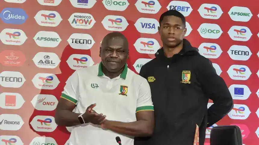 L'entraîneur et le joueur camerounais posent devant un fond de sponsor lors d'une conférence de presse, affichant la fierté de l'équipe avant un tournoi.