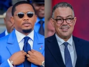 Samuel Eto'o en costume bleu ajustant sa cravate, aux côtés de Fouzi Lekjaa en costume sombre avec des lunettes, tous deux souriant sur un fond flou.