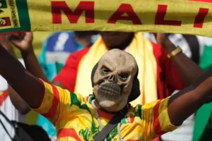Un fan malien en maillot vibrant agite une écharpe jaune "MALI", portant un masque de crâne distinctif, montrant l'esprit d'équipe pendant un match.