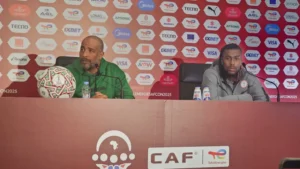 Entraîneur et joueur marocains lors d'une conférence de presse, discutant des prochains matchs avec un fond de marque et un ballon de football sur la table.
