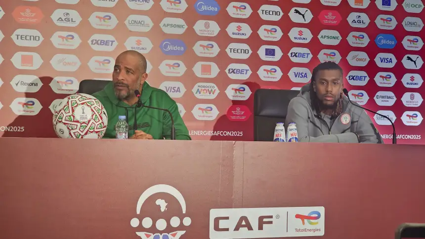 Entraîneur et joueur marocains lors d'une conférence de presse, discutant des prochains matchs avec un fond de marque et un ballon de football sur la table.