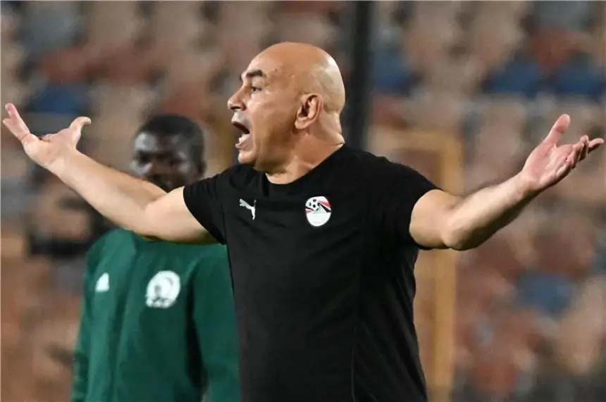 Coach passionately gestures during a match, wearing a black shirt with Egyptian flag crest, with another staff member in green behind.