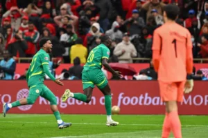 Senegal players celebrate a goal in vibrant green kits, with a cheering crowd in the background and a goalkeeper in orange.