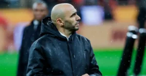 Walid Regragui, Morocco's national team coach, stands in the rain, focused and thoughtful, wearing a dark jacket, with a blurred stadium background.