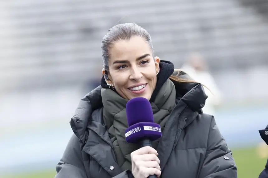 Un présentateur sportif sourit en tenant un micro gainé de violet, vêtu d'une veste vert foncé sur fond de stade flou.
