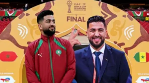 Morocco player stands confidently beside an organiser at the AFCON 25 final backdrop, featuring vibrant colours and national symbols.