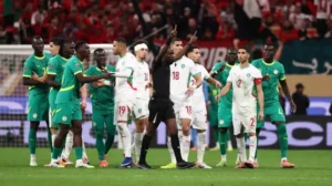 Morocco and Senegal players argue with the referee during a tense moment in their international match, showcasing vibrant team kits and stadium atmosphere.