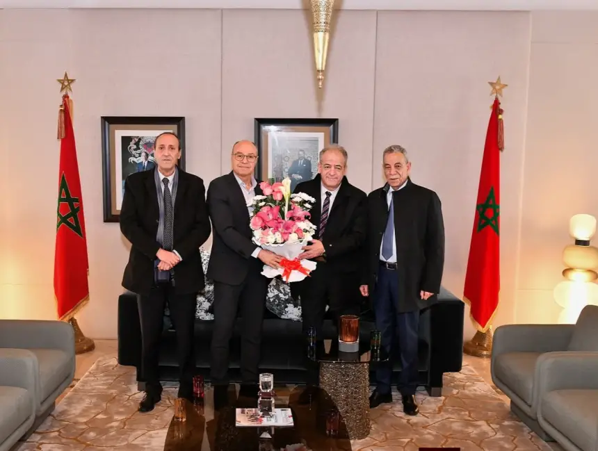 Four men pose together in a room adorned with Moroccan flags, holding a bouquet of pink flowers, symbolising celebration and unity.