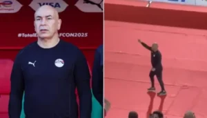 Hossam Hassan, gekleed in het teamoutfit van Egypte, staat voor een rode achtergrond van het veld en gebaart tijdens een wedstrijd tegen Benin op AFCON 2025.