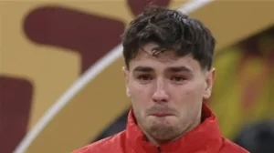Un joueur de football en maillot rouge en pleurs, visiblement ému pendant un match, avec un fond de stade flouté mettant en valeur l'atmosphère.