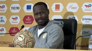 Senegal's football coach at a press conference, looking thoughtfully at a patterned match ball, against a bright yellow backdrop featuring sponsors.