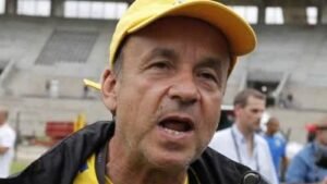 Entraîneur parlant avec passion, coiffé d'une casquette jaune ; l'arrière-plan du stade évoque un match récent ou une séance d'entraînement.