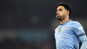 Joueur de Manchester City en tenue bleu clair, ayant l'air pensif pendant un match, avec le badge FIFA visible sur la poitrine.