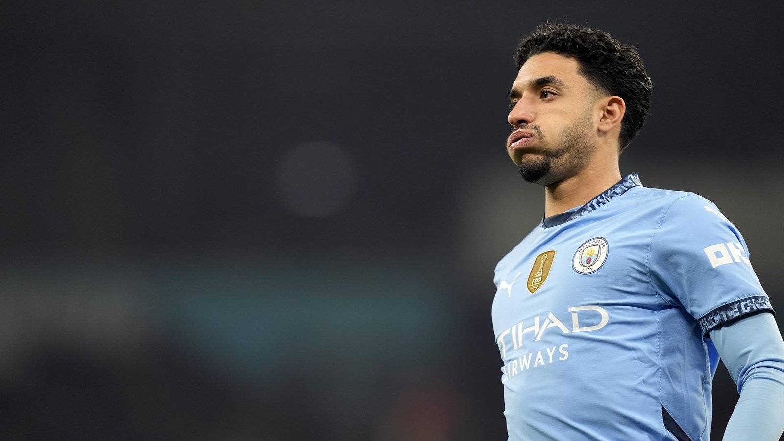 Joueur de Manchester City en tenue bleu clair, ayant l'air pensif pendant un match, avec le badge FIFA visible sur la poitrine.