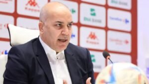 Moroccan football official passionately speaks at a press conference, seated in front of branded sponsor logos, with a football on the table.