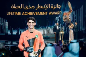 Yassine Bounou in a bright orange goalkeeper kit holds a trophy at a ceremony backdrop featuring "Lifetime Achievement Award" in Arabic and English.