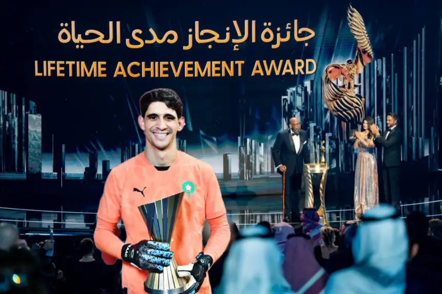 Yassine Bounou in a bright orange goalkeeper kit holds a trophy at a ceremony backdrop featuring "Lifetime Achievement Award" in Arabic and English.