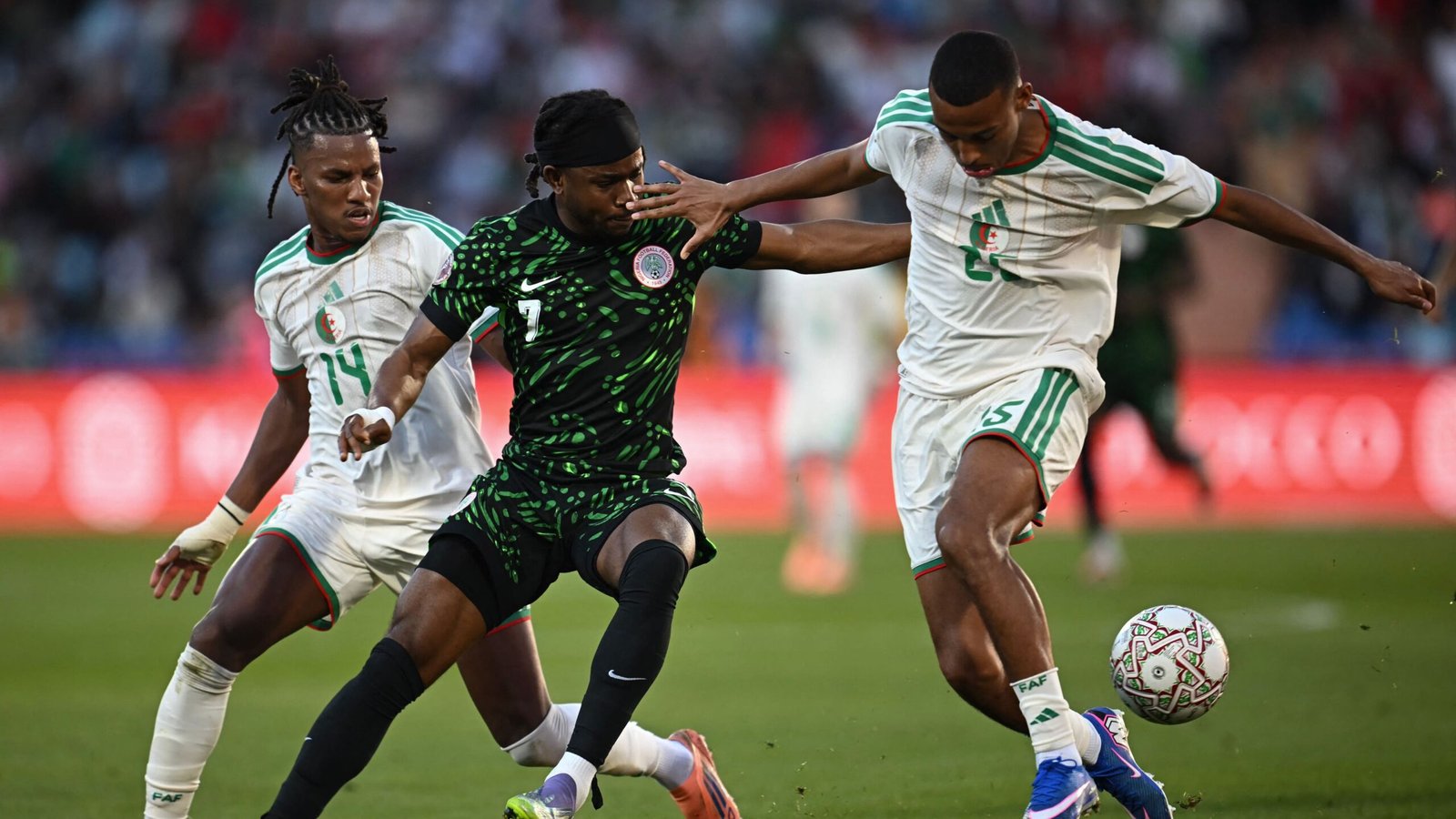 Un joueur nigérian en maillot vert lutte contre des adversaires algériens en blanc pendant un moment tendu du match, avec le ballon en jeu.