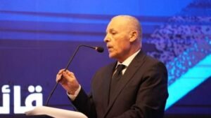 Moroccan football official delivering a speech at an event, dressed in a dark suit, with a microphone and a blue themed background.
