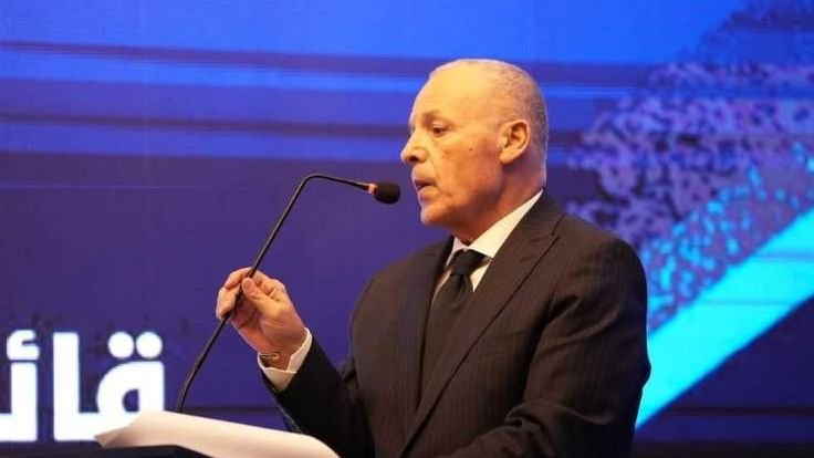 Moroccan football official delivering a speech at an event, dressed in a dark suit, with a microphone and a blue themed background.