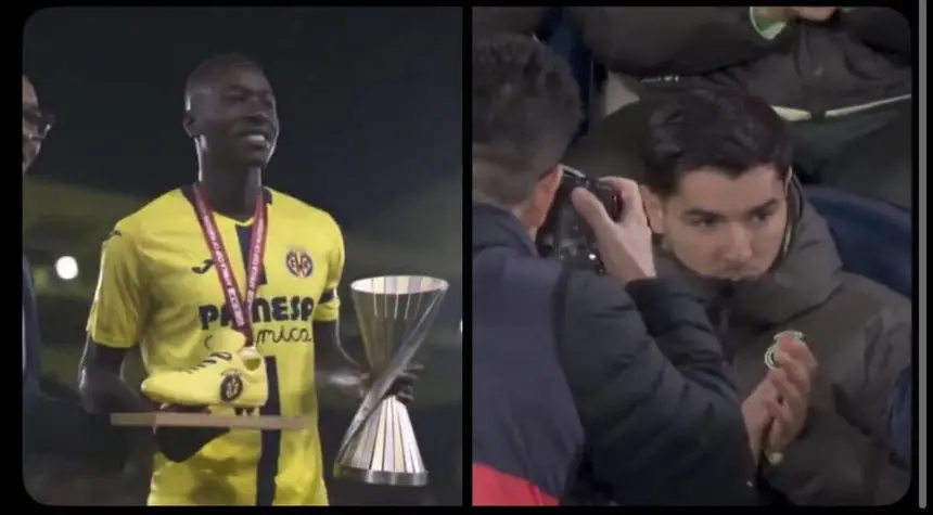 Doelman Yassine Bounou viert met een trofee in het gele tenue van Villarreal, terwijl een fan op de tribune applaudisseert en foto's maakt.
