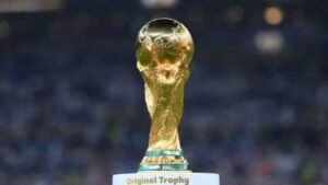Golden FIFA World Cup trophy displayed on a pedestal with "Original Trophy" label, surrounded by a blurred stadium backdrop.