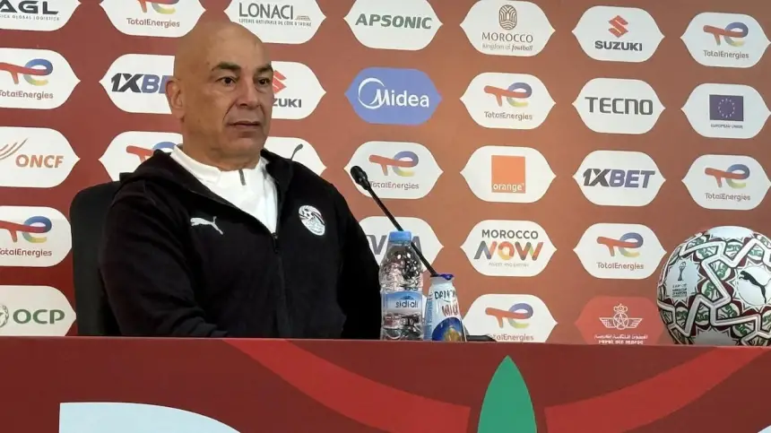 L'entraîneur assis lors d'une conférence de presse avec un fond rouge et blanc arborant des logos de sponsors, un ballon de football à ses côtés, se concentrant sur des discussions concernant le football marocain.