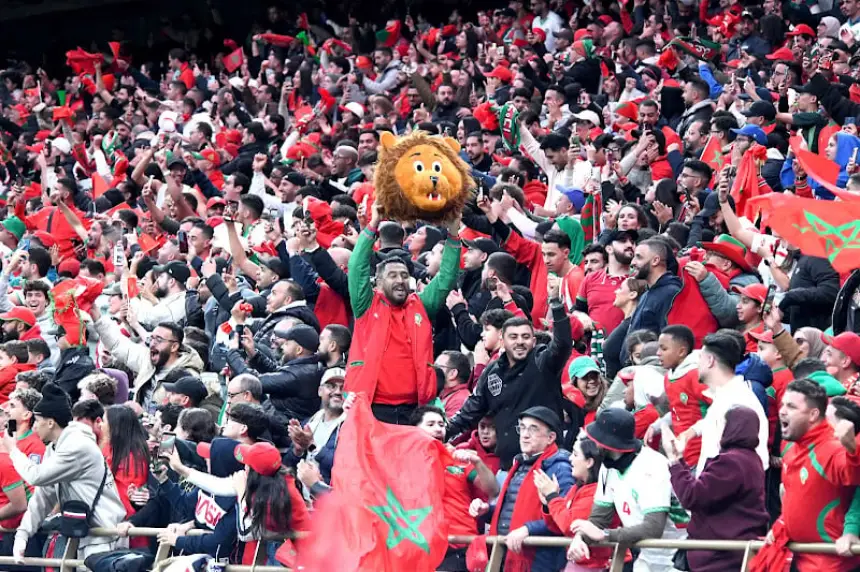 Les supporters marocains célèbrent avec passion, vêtus de rouge et de vert, avec des drapeaux agités et un supporter brandissant une tête de mascotte lion.