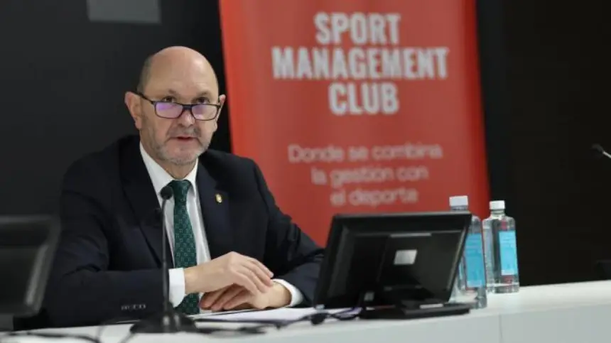 رجل يرتدي بدلة داكنة ونظارات يتحدث أمام طاولة تحمل حاسوباً، وتحيط به قناني ماء، أمام لافتة حمراء لنادي إدارة الرياضة.