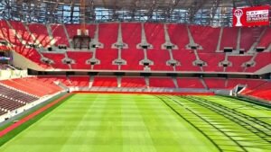 داخل ملعب فارغ يتميز بمقاعد حمراء وملعب أخضر مهيأ جيدًا، يعرض شعار كأس الأمم الأفريقية CAF المغرب 2025.