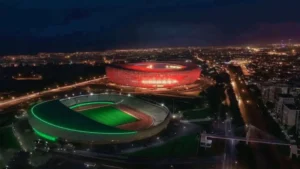 Vue aérienne nocturne de deux stades ; le stade supérieur brille en rouge tandis que l'inférieur présente des éclats verts vibrants, sur fond de paysage urbain.