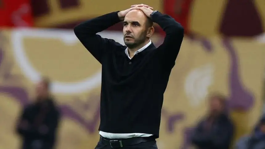 مدرب كرة قدم مغربي يبدو متضايقًا واضعًا يديه على رأسه، مرتديًا سترة سوداء. خلفية الملعب توحي بلحظة مباراة مشحونة.