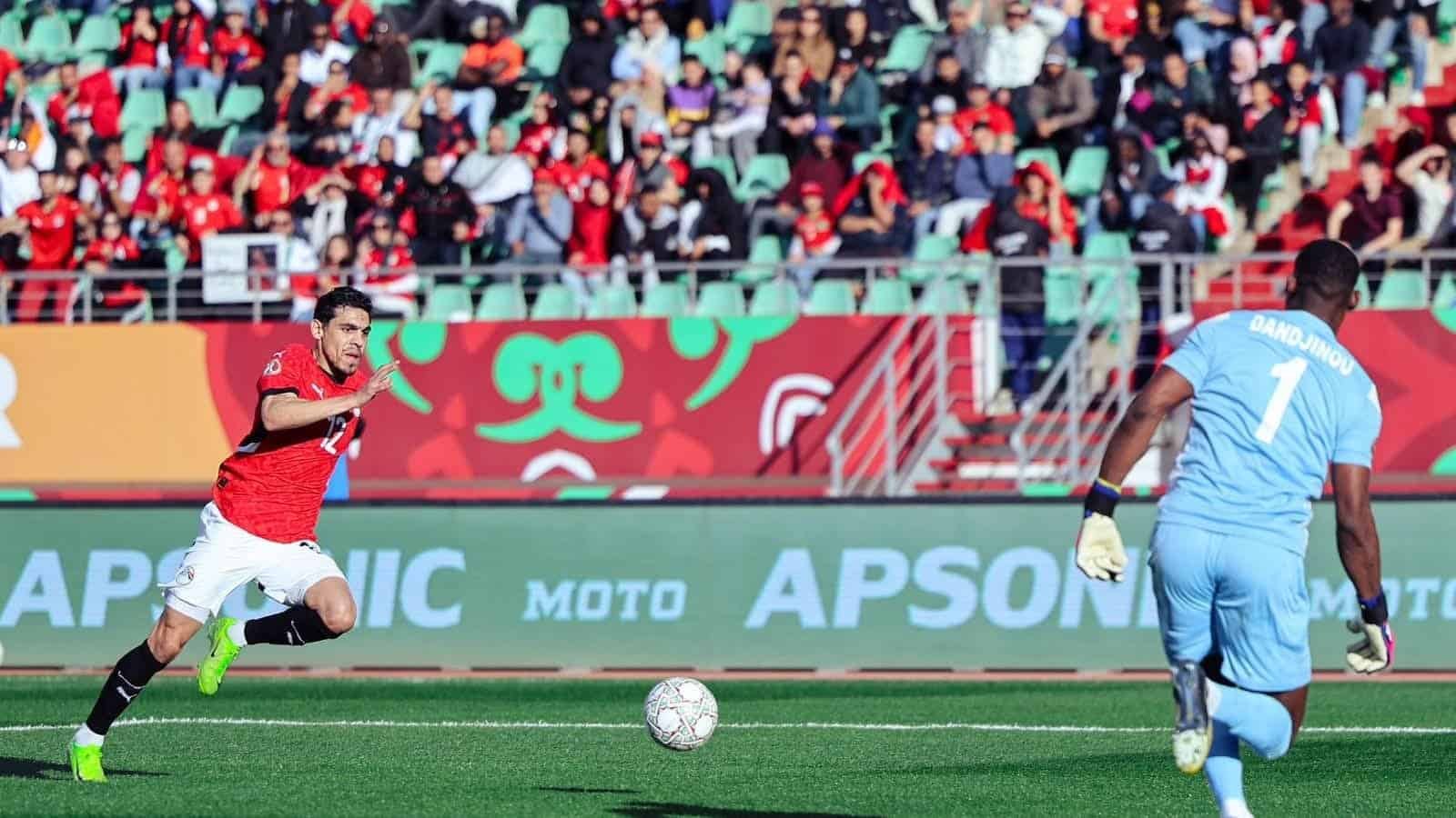 Un joueur marocain en tenue rouge sprinte vers le but, tandis que le gardien adverse en bleu clair se prépare à défendre, avec des supporters en arrière-plan.