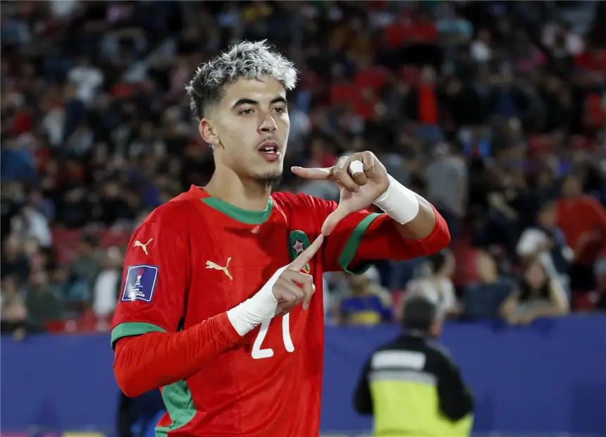 Un footballeur marocain en tenue rouge célèbre, effectuant un geste des mains, au sein d'une foule enthousiaste dans un stade pendant un match.