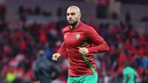 Marokkaanse speler draagt een rood trainingsshirt met groene accenten, warmt op op het veld te midden van een juichende menigte in het stadion.