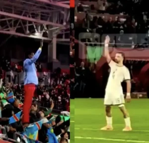 Un supporter en veste bleue et pantalon rouge agite énergiquement les bras, entouré de partisans brandissant des drapeaux, aux côtés d'un joueur en tenue blanche qui salue.