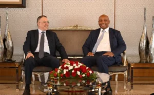 Two men seated comfortably on a plush sofa in a well-decorated room, smiling and engaged in conversation, with a table adorned with red roses in front.