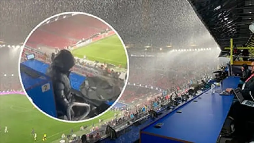 Zware regen valt op een voetbalstadion, met op de voorgrond een toeschouwer in een donkere jas met capuchon en zichtbare lege zitplaatsen.