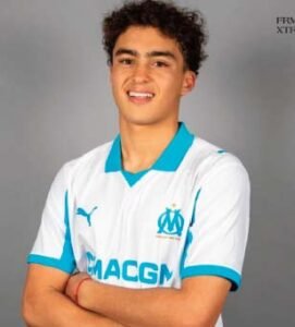 Young Moroccan player wearing Olympique Marseille's white kit with turquoise accents, smiling confidently and posing with crossed arms.