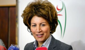 Moroccan football official with curly hair speaking to reporters, background features a stylised football logo in green and red.