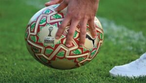 Hand gripping a colourful ball designed for the Africa Cup of Nations, set against a green football pitch backdrop.