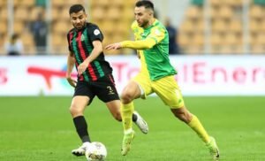 Twee voetballers strijden om de bal; één in een rood-groen gestreept tenue en zwarte shorts, de ander in een geel tenue met groene accenten.