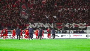 Celebrating players in red jerseys acknowledge passionate fans holding vibrant banners and flags, displaying "Winners" in a festive stadium atmosphere.
