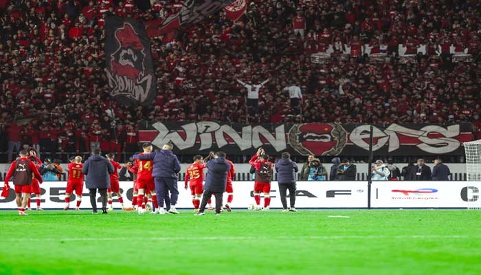 Celebrating players in red jerseys acknowledge passionate fans holding vibrant banners and flags, displaying "Winners" in a festive stadium atmosphere.